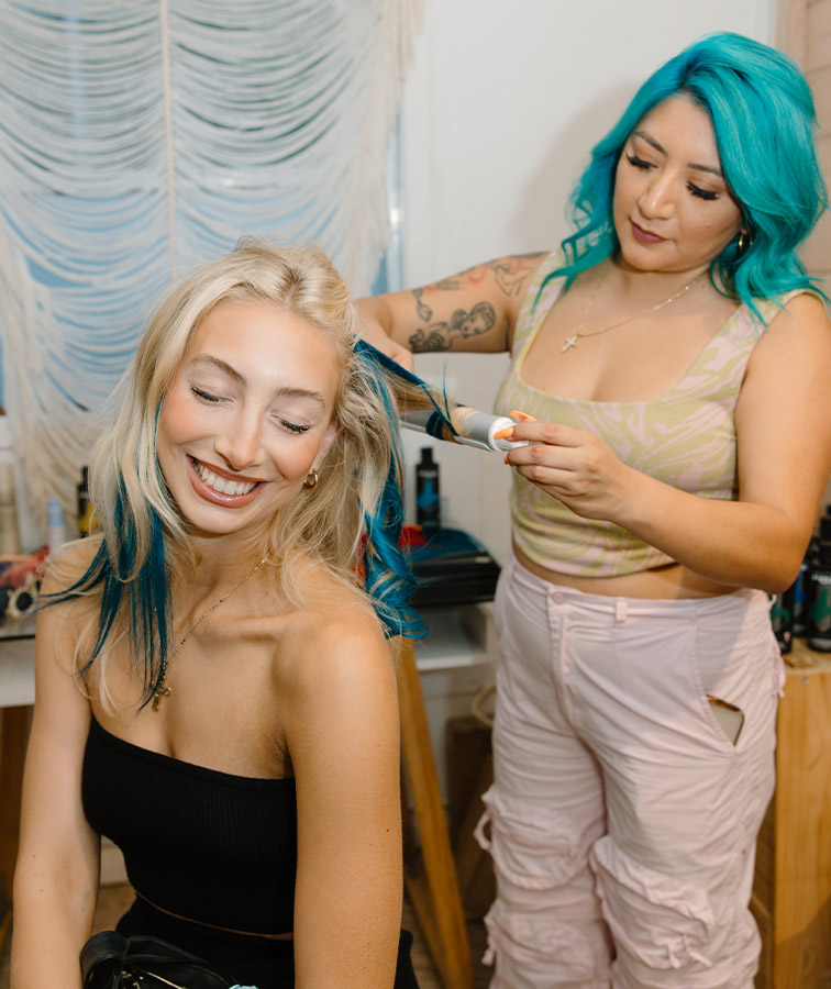 A cheerful young woman with blonde hair laughs while receiving a vibrant blue hair dye treatment from a stylist with striking teal hair, in a creatively decorated salon setting.